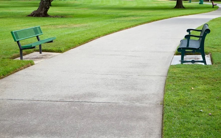 Palm Beach Concrete. A green bench rests on the grass beside a paved pathway, inviting relaxation in a serene outdoor setting.